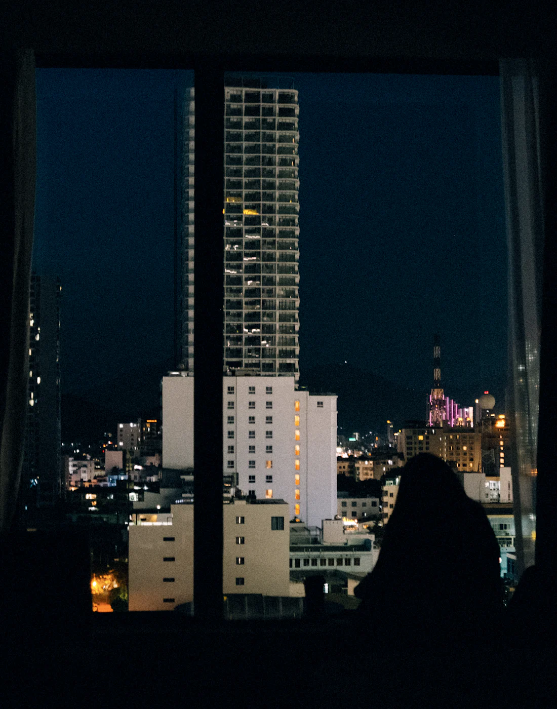 A woman in silhouette at a window overlooking the city at night